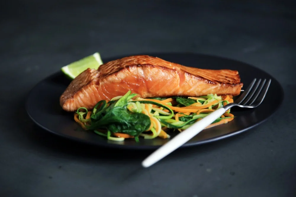 A seared salmon fillet on a dark plate over zucchini and carrot noodles with spinach, with a lime wedge and a fork.